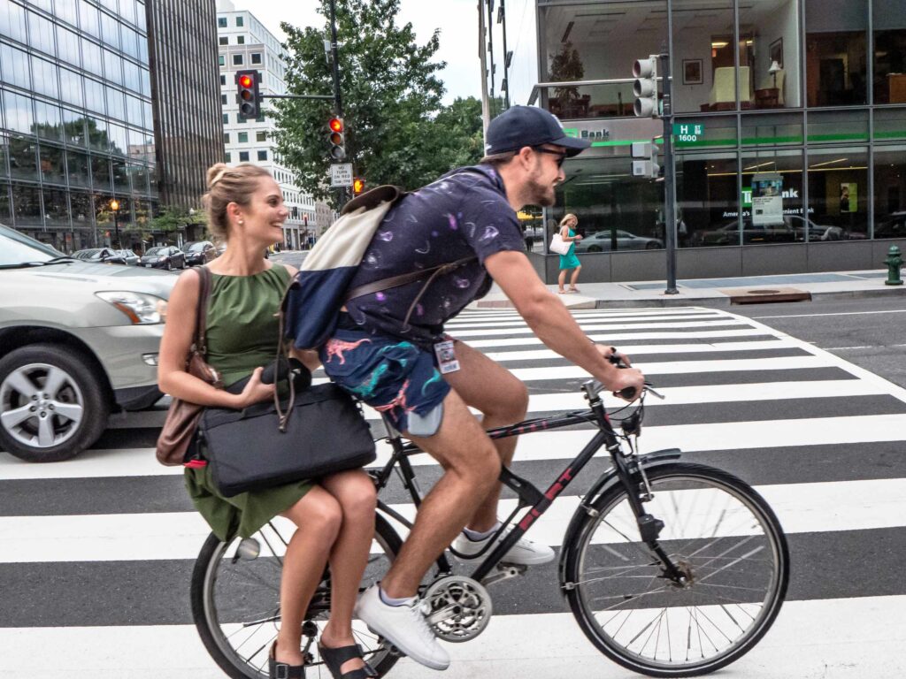 A young man and women pass by on a bike, the man peddling and the smiling woman sitting on the small bike rack behind him.