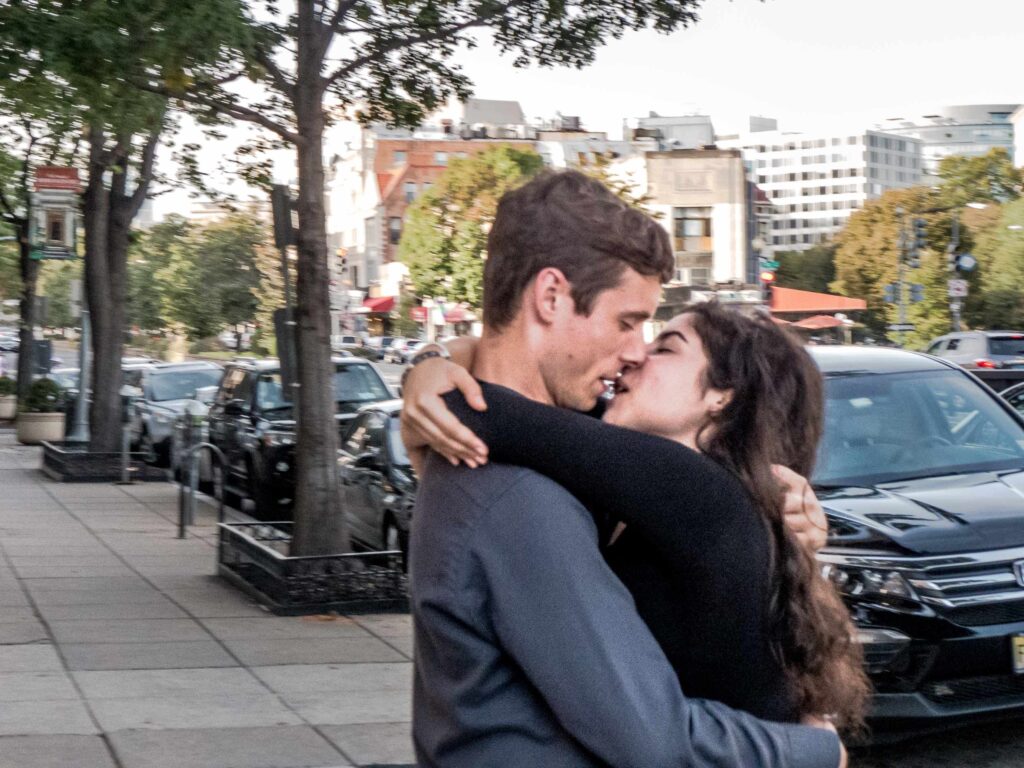 A couple standing on a sidewalk, arms locked around each other and eyes closed in the moment their mouths are about to lock in the embrace of a passionate kiss.