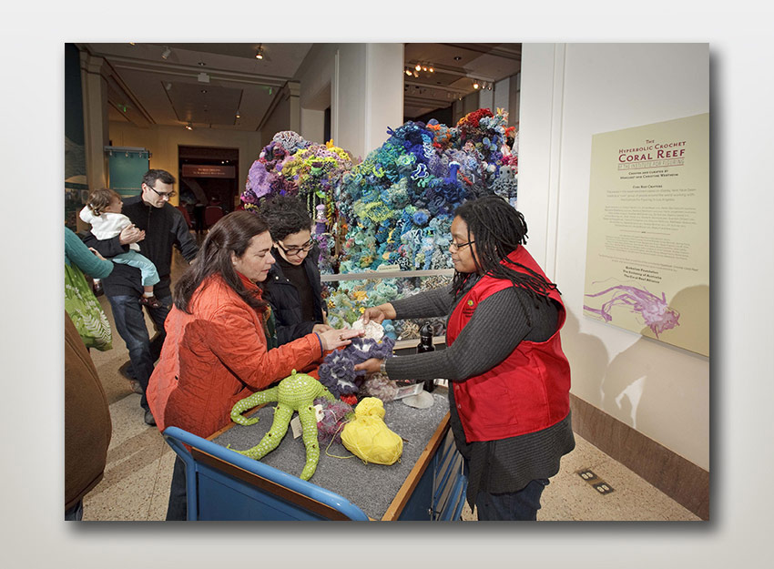 Volunteer for the exhibit,  "Hyperbolic Crochet Coral Reef" showing visitors details of some ocean organisms and crocheted items at an activity cart situated in the exhibit.